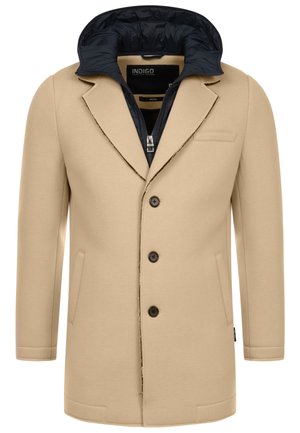 Beige ytterjakke med marineblå innerfôr, laget av glatt stoff. Har to sidelommer, en enkelt brystlomme og svarte knapper.