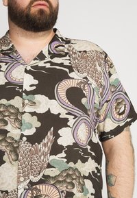 Lyhythihainen paita tummanruskeasta kankaasta, jossa on yksityiskohtainen kuvio lintuista, käärmeistä ja pilvistä hillityissä väreissä. Napitettava malli.