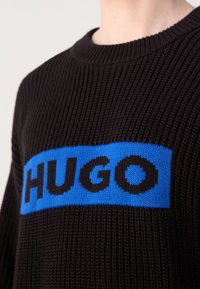 Pull en maille côtelée noire avec un patch rectangulaire bleu affichant le texte "HUGO" en lettres noires en gras sur la poitrine.
