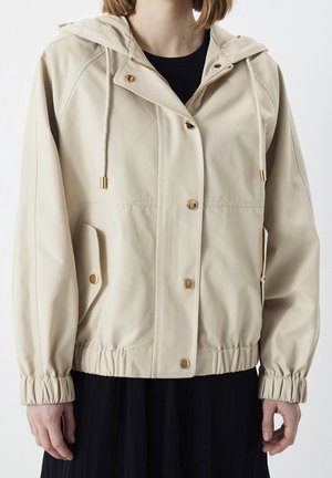 Personne portant une veste beige à capuche avec des boutons dorés, un devant matelassé, des cordons, des poignets élastiques et des poches à rabat.