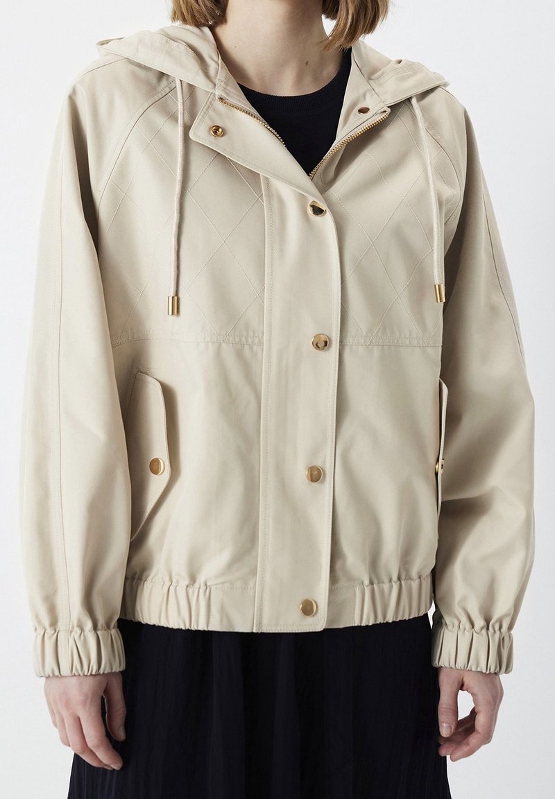 Personne portant une veste beige à capuche avec des boutons dorés, un devant matelassé, des cordons, des poignets élastiques et des poches à rabat.