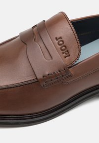 Mocassin en cuir marron avec semelle noire et logo "JOOP!" en relief sur la sangle supérieure, avec des coutures détaillées sur fond blanc.