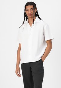 Short-sleeve white shirt in lightweight fabric, featuring a button-down front, a classic collar, and a relaxed fit. Paired with dark trousers.