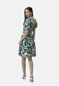 Blumenmusterkleid in Grün, Rot und Weiß. Puffärmel, quadratischer Rücken, tailliert und knielang. Tragen mit weißen Absätzen.
