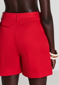 Primo piano di una persona che indossa pantaloni corti su misura di un vivace rosso, con tasche posteriori, mostrando la parte centrale della schiena e un braccio con bracciali bianchi e marroni.