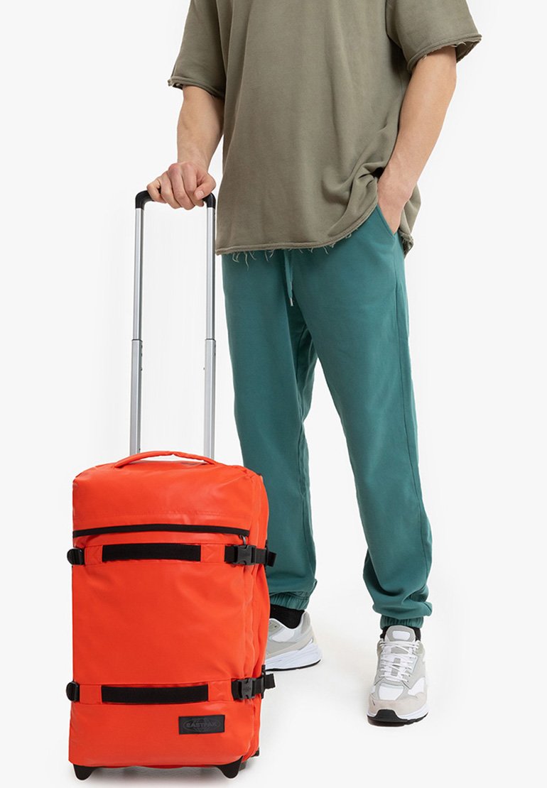 Bolsa de deporte naranja con ruedas, con correas y herrajes negros, parte superior redondeada, tela resistente y un bolsillo con cremallera en la parte frontal; sostenida por una persona vestida con ropa casual.