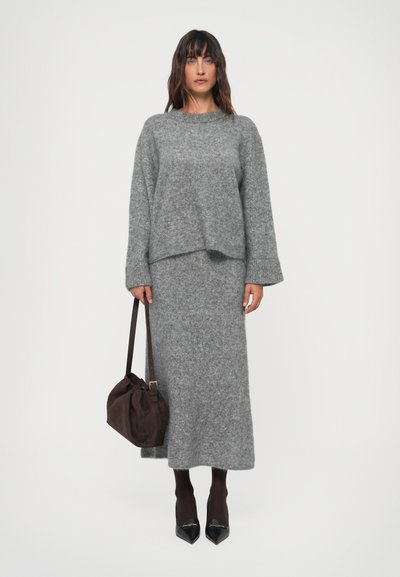 Maglione grigio a maglia con spalle scese, abbinato a una lunga gonna coordinata. La figura tiene una borsa in suede marrone e indossa tacchi neri.