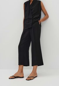 Haut noir sans manches à nouer, associé à un pantalon noir à taille haute, jambes larges et longueur courte, et des sandales plates noires, debout contre un mur blanc.