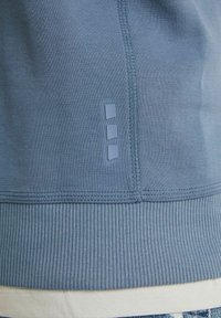 Modrá mikina s ribbovaným lemem, se třemi malými modrými obdélníkovými akcenty po straně. Hladká textura a přiléhavý střih.