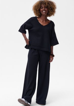 Femme aux cheveux bouclés souriante, portant un top noir ample à col en V et un pantalon noir large, se tenant contre un fond blanc.