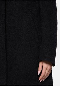 Zwarte textuurjack met een zachte, fuzzy stof. Heeft een losse pasvorm, lange mouwen en zijzakken. De hand die op de stof rust, toont een neutraal nageldesign.
