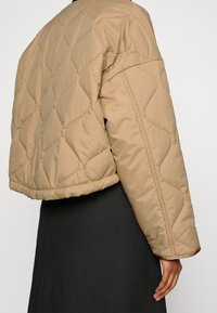 Person iført en quilted, lys brun cropped jakke over en lang, sort nederdel, med hånden afslappet ved siden af.