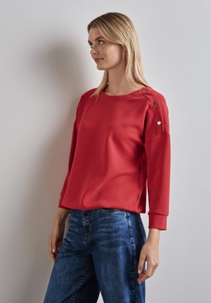 Rotes leichtgewichtiges Sweatshirt mit langen Ärmeln, rundem Ausschnitt und dekorativen Knöpfen an der Schulter. Kombiniert mit blauen Jeans.
