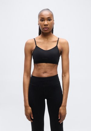 Mujer atlética con el cabello recogido, vestida con un sujetador deportivo negro y leggings negros de talle alto, posando contra un fondo gris claro liso.