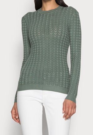 Femme portant un pull en tricot texturé à manches longues de couleur vert sauge avec un motif en chevron et un pantalon blanc, se tenant devant un fond uni.