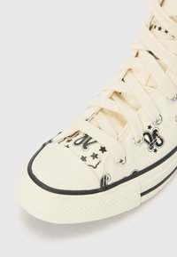 Leinenschuhe mit cremefarbenem Hintergrund und schwarzen Grafikdrucken, darunter Sterne und Buchstaben. Mit weißen Schnürsenkeln und Gummisohle ausgestattet.