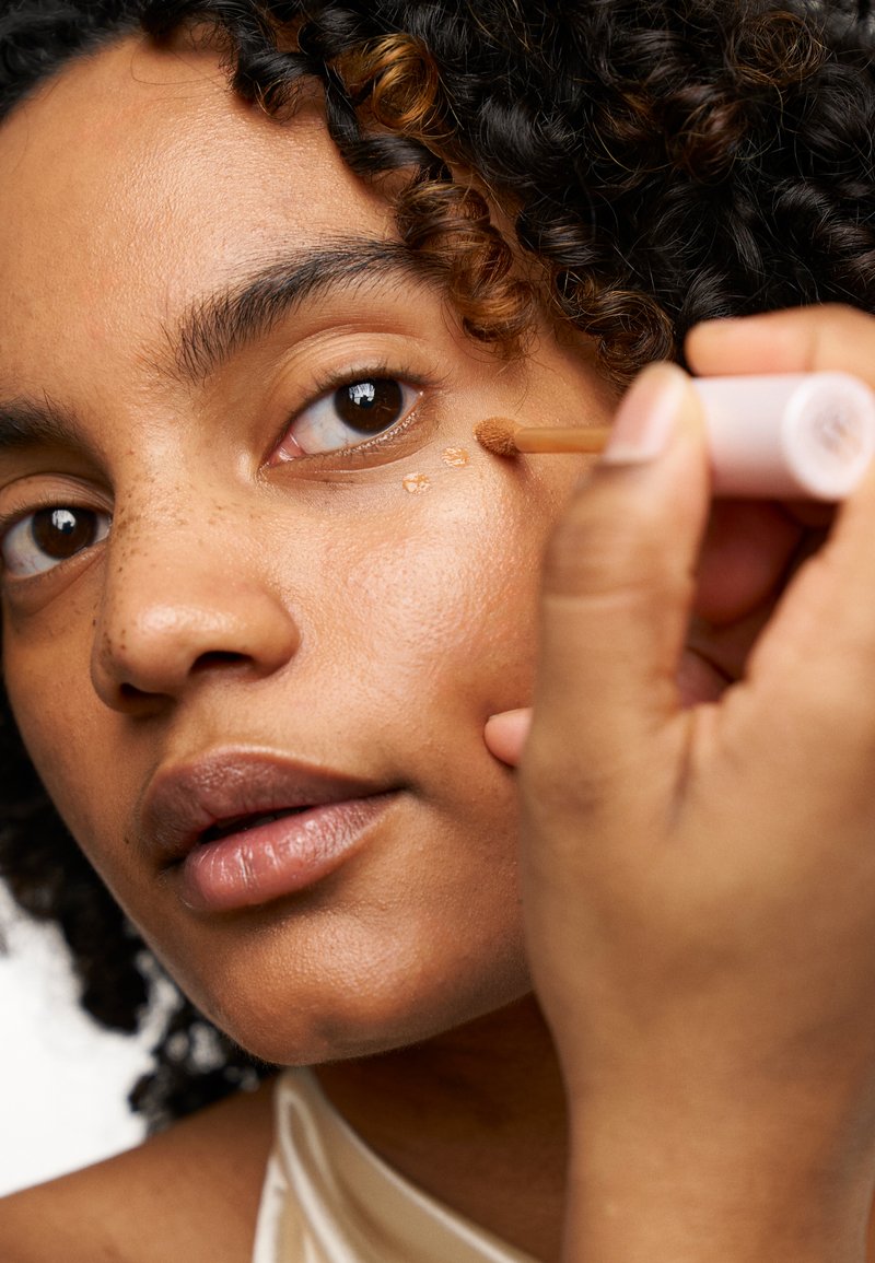 Un correcteur liquide est appliqué sous un œil marron ; le modèle a des cheveux frisés. La peau montre une texture naturelle avec des petites taches de rousseur visibles.