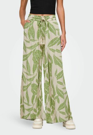 Kvinde iført beige bukser med brede ben og grønt bladprint, snøre i taljen, kombineret med hvide sneakers og sort ærmeløs top.