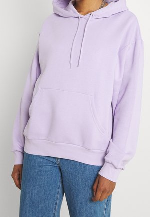 Person iført lyslilla hættetrøje med frontlomme og snore, kombineret med blå jeans, vist fra nakke til hofter.