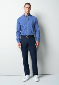 Lichtblauw overhemd met knopen, marineblauwe broek, witte sneakers. Het overhemd heeft een kraag en een geborduurd logo. De broek heeft een gestructureerd patroon.