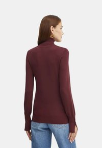 Maglione a collo alto bordeaux con polsini e orlo a costine, caratterizzato da un design aderente e una superficie liscia. Indossato con jeans larghi azzurri.
