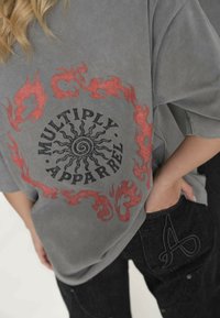 Szary oversize t-shirt z czerwonymi grafikami płomieni oraz napisem "MULTIPLY APPAREL". Noszony z ciemnymi dżinsami, z logo na kieszeni.