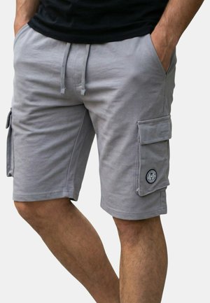 Homme portant un short cargo gris clair avec poches latérales et un cordon noir, les mains dans les poches, debout devant un fond uni.