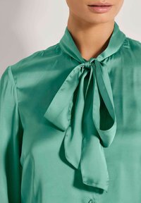 Grüne Seidenbluse mit einer gebundenen Schleife am Kragen, glatter Textur und Knopfverschluss. Der Stoff hat eine glänzende Oberfläche.