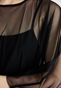 Black sheer top with a translucent design, featuring layered, flowing sleeves and a structured neckline, creating a contrast with skin tone.