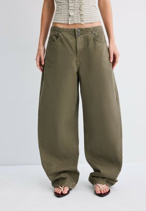 Personne portant un pantalon ample vert olive, un haut court à carreaux et des sandales blanches en forme de tongs, debout devant un fond uni.
