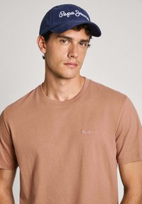 Camiseta de algodón marrón de mangas cortas con un pequeño logo blanco de "Pepe Jeans" en el lado izquierdo del pecho. Gorro azul con bordado de logo blanco.