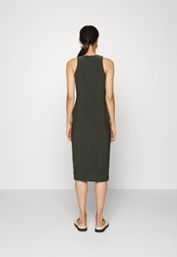 Žena s tmavým culíkem, oblečená v bezramínkových tmavozelených midi šatech a bílých pantoflích, stojící zády k jednoduchému pozadí.