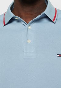 Polo bleu clair en tissu texturé, avec un accent rouge sur le col, trois boutons, et un petit logo dans un coin aux couleurs rouge, blanc et bleu.