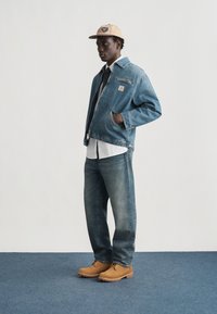 Άνδρας με τζιν μπουφάν, τζιν παντελόνι, μπεζ καπέλο, λευκό πουκάμισο, μαύρη γραβάτα και καφέ μπότες εργασίας, στέκεται σε μπλε δάπεδο μπροστά σε λευκό τοίχο.