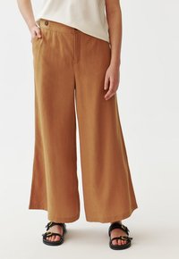 Personne portant un pantalon marron à taille haute et jambes larges, un haut crème rentré à l'intérieur, et des sandales noires avec des boucles dorées, se tenant sur un fond blanc.