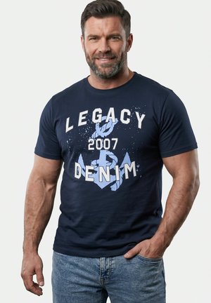 Brodaty mężczyzna w granatowej koszulce z wzorem "Legacy 2007 Denim" i kotwicą, noszący jasnoniebieskie dżinsy, uśmiecha się z jedną ręką w kieszeni.