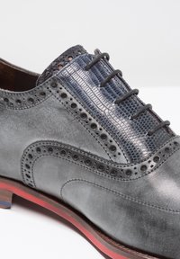 Scarpa in pelle grigia con dettagli brogue, accentuazione in pelle blu testurizzata e suola con riflessi rossi. Presenta punta arrotondata e lacci neri.
