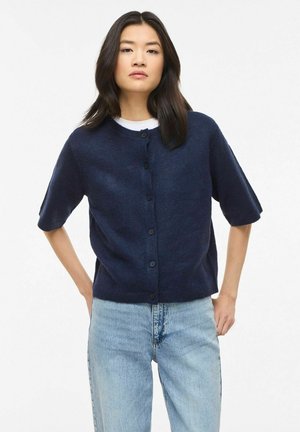 Fiatal nő egyenes sötét hajjal sötétkék, gombos rövid ujjú kardigánt és világoskék, magas derekú farmert visel.