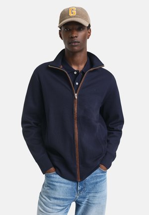 Marineblaues Zip-Up Sweatshirt mit braunen Akzenten, weichem Stoff und hohem Kragen. Über einem Poloshirt getragen, ergänzt durch eine beige Kappe mit einem Logo.