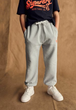 Pantaloni tuta grigi con vita elastica e cordino bianco, abbinati a una maglietta nera con testo rosso e sneakers bianche.