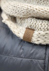 Sciarpa di colore crema lavorata a maglia con un motivo testurizzato, con un'etichetta in pelle marrone, posata su un tessuto di giacca imbottita grigio scuro.