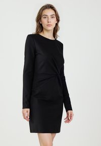 Czarna sukienka z długim rękawem wykonana z gładkiej tkaniny, z zawiązanym węzłem w talii i długością do kolan. Czysta, minimalistyczna sylwetka.