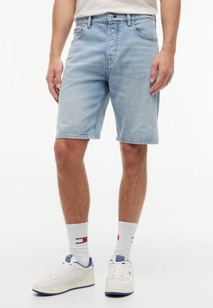 Hele sinised teksashortsid, mida kannab seisvas asendis inimene, koos valgete sokkidega, millel on punase, valge ja sinise logoga ning valgete tossudega.