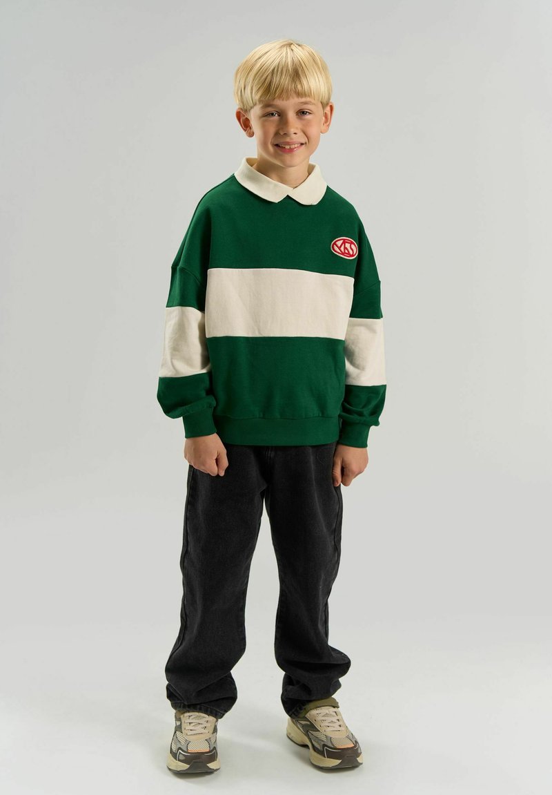 Enfant blond debout, portant un sweat à capuche rayé vert et blanc, un pantalon noir et des baskets beiges, souriant sur un fond uni.