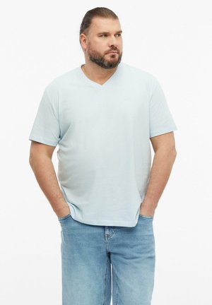 Mężczyzna z brodą i związanymi włosami, ubrany w jasnoniebieski T-shirt z dekoltem w serek i niebieskie dżinsy, z rękami w kieszeniach, patrzący na bok.