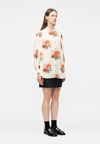 Γυναίκα που φοράει μακρυμάνικο εκρού πουκάμισο με πορτοκαλί λουλουδάτο μοτίβο, μαύρη δερμάτινη μίνι φούστα, μαύρες κάλτσες και μαύρα μοκασίνια, στέκεται πλευρικά.