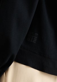 Black ribbed sweatshirt with a smooth texture; features a black logo print near the hem. Paired with a light beige fabric below.