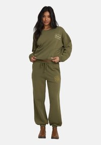 Kobieta nosząca oliwkowo-zieloną bluzę z logo fali i dopasowane dresy z motywem słońca, do których założone są brązowe botki za kostkę.