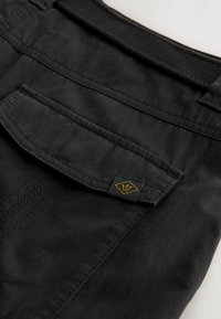 Poche de pantalon noire avec rabat et petite étiquette arborant un logo trident jaune, montrant les coutures détaillées et la texture du tissu.
