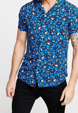 Mężczyzna w niebieskiej koszuli z krótkim rękawem z geometrycznym wzorem i czarnych spodniach, pokazujący tatuaż na prawej ręce.